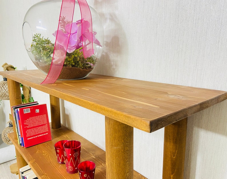 May include: A wooden shelf unit with two shelves. The top shelf has a glass globe with pink flowers and greenery inside. The bottom shelf has a stack of books and three red candle holders.