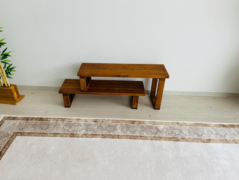 K&ouml;nnte beinhalten: Ein zweistufiger Holztisch mit einer nat&uuml;rlichen braunen Oberfl&auml;che. Die obere Stufe ist breiter als die untere Stufe und beide haben eine einfache rechteckige Form.