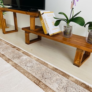 K&ouml;nnte beinhalten: Ein TV-St&auml;nder aus Holz mit zwei Regalen. Das obere Regal hat einen Fernseher darauf und das untere Regal hat drei B&uuml;cher mit den Worten "Coco Chanel" auf dem Einband. Auf dem St&auml;nder stehen zwei Topfpflanzen, eine mit lila Bl&uuml;ten und eine mit gr&uuml;nen Bl&auml;ttern.