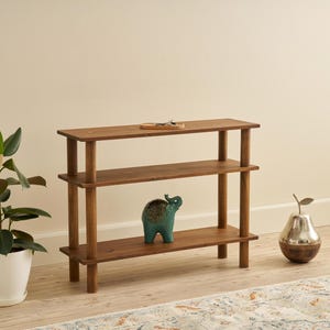 Puede incluir: Una estantería de madera de tres niveles con acabado marrón oscuro. Una pequeña figura de elefante turquesa se encuentra en el estante del medio. Una planta en maceta y un objeto decorativo en forma de pera también están en la imagen.