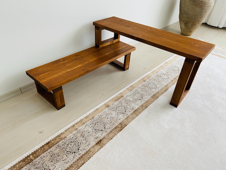 K&ouml;nnte beinhalten: Eine Holzbank und ein l&auml;ngerer Holztisch mit einer nat&uuml;rlichen braunen Oberfl&auml;che. Tisch und Bank stehen auf einem wei&szlig;en Teppich mit braunem Muster an der Kante.