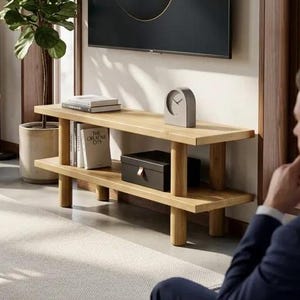 May include: A light-coloured wooden media console with two shelves. The top shelf holds books and a grey clock. The bottom shelf has a black box. A large TV is mounted on the wall above the console.