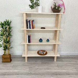 May include: A natural wood bookshelf with four shelves. The shelves are empty except for a few books, a small bowl, and two decorative items. The bookshelf is standing on a light gray floor.