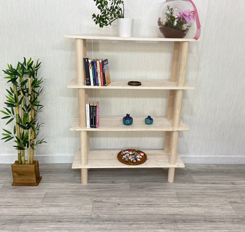 May include: A natural wood bookshelf with four shelves. The shelves are empty except for a few books, a small bowl, and two decorative items. The bookshelf is standing on a light gray floor.