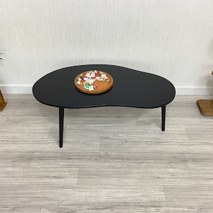 May include: A black, kidney-shaped coffee table with four slender legs. A wooden tray with small decorative items sits on the table's surface. The table is in a room with light-colored flooring and a white wall.