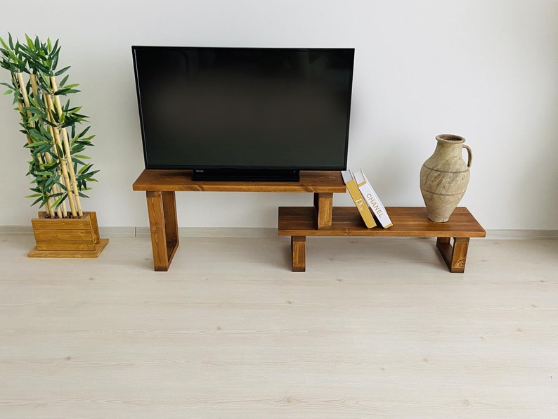 K&ouml;nnte beinhalten: Ein TV-St&auml;nder aus Holz mit zwei Regalen. Das obere Regal h&auml;lt einen Flachbildfernseher und das untere Regal eine Vase und zwei B&uuml;cher. Der St&auml;nder ist aus dunkelbraunem Holz und hat einen rustikalen Look.