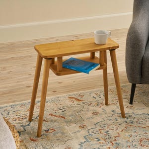 May include: A small, light-brown wooden side table with a rectangular top and a lower shelf. A white mug and a blue book rest on the table. The table is supported by four angled legs. The table is 24 inches tall.