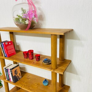May include: A wooden shelf unit with four shelves. The shelves are made of brown wood and have a rustic look. The shelf unit is empty except for a few books, a glass vase with pink flowers, and some red candle holders.