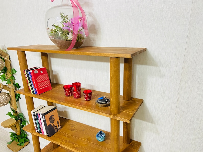 May include: A wooden shelf unit with four shelves. The shelves are made of brown wood and have a rustic look. The shelf unit is empty except for a few books, a glass vase with pink flowers, and some red candle holders.