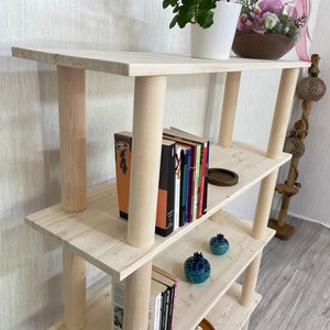 May include: A light wood bookshelf with four shelves. The shelves are stacked on top of each other and are supported by four wooden posts. There are books, decorative items, and a plant on the shelves.