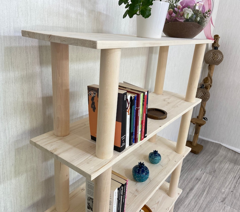 May include: A light wood bookshelf with four shelves. The shelves are stacked on top of each other and are supported by four wooden posts. There are books, decorative items, and a plant on the shelves.