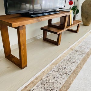 K&ouml;nnte beinhalten: Ein TV-St&auml;nder aus Holz mit zwei Ablagen. Der St&auml;nder ist aus dunkelbraunem Holz und hat eine rechteckige Form. Der St&auml;nder steht auf einem wei&szlig;en Teppich mit braunem Muster.