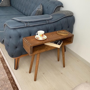 May include: A wooden side table with a shelf and four legs. The table is stained a dark brown color and has a rustic look. The table is next to a blue sofa and has a cup and saucer, a book, and a coaster on top.