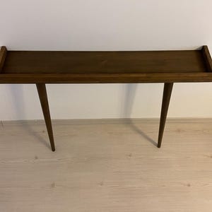 May include: A dark brown wooden console table with a rectangular top and two slender legs. The table is standing on a light wood floor.