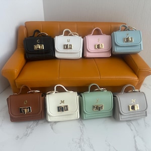 Peut inclure: Collection de huit mini-sacs à main de différentes couleurs, dont noir, blanc, rose et bleu. Chaque sac est doté d'un fermoir et d'une poignée dorés, avec une lettre sur le devant. Les sacs sont disposés sur un petit canapé orange.