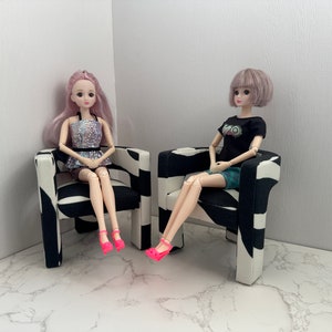 May include: Two dolls sitting in black and white patterned chairs. The doll on the left has pink hair and is wearing a silver sequined top and a black skirt. The doll on the right has purple hair and is wearing a black shirt with a graphic print and a green skirt.