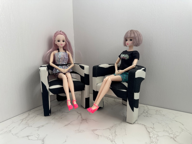 May include: Two dolls sitting in black and white patterned chairs. The doll on the left has pink hair and is wearing a silver sequined top and a black skirt. The doll on the right has purple hair and is wearing a black shirt with a graphic print and a green skirt.