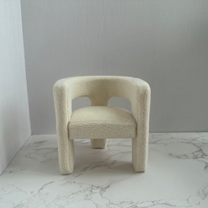 May include: A white plush armchair with a rounded back and a simple design. The chair is sitting on a white marble surface.