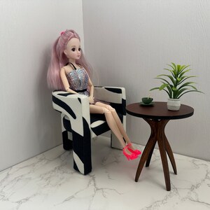 May include: A doll sitting in a zebra-print armchair with a small wooden table next to it. The table has a green plant in a white pot and a small green dish on top.