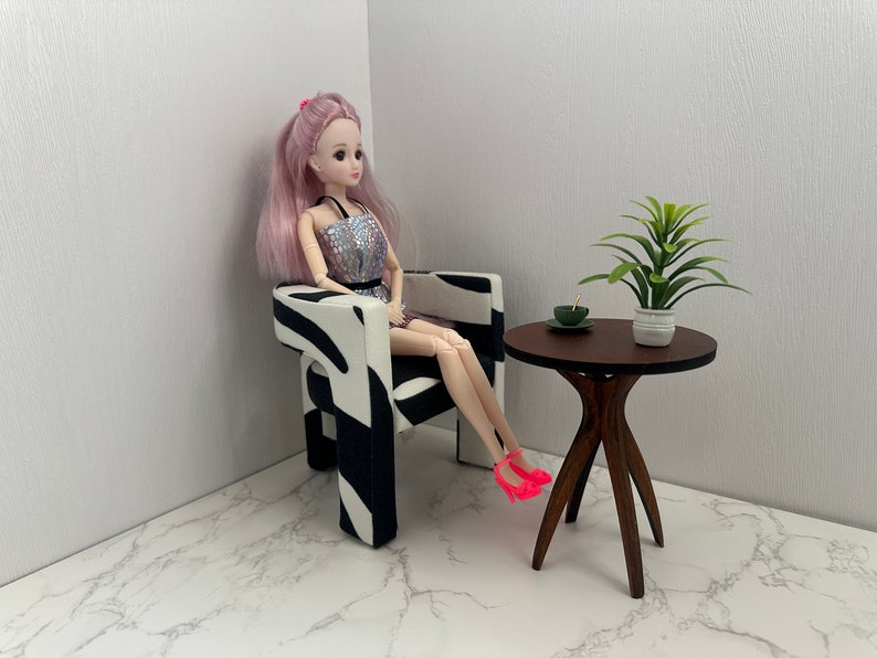 May include: A doll sitting in a zebra-print armchair with a small wooden table next to it. The table has a green plant in a white pot and a small green dish on top.