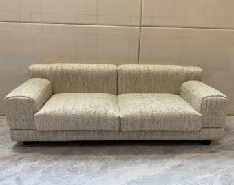 May include: A light beige fabric sofa with a tufted back and armrests. The sofa has a simple, modern design and is perfect for a small living room or apartment.