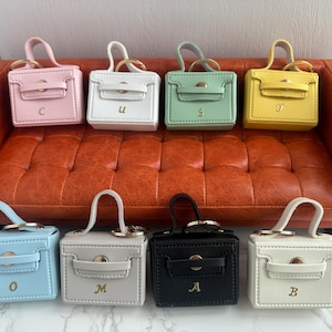 Puede incluir: Una colección de pequeños bolsos cuadrados en varios colores pastel, cada uno con un asa superior y un cierre dorado. Las bolsas están dispuestas sobre un sofá naranja. Cada bolso tiene una letra.
