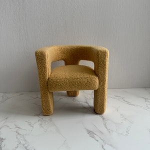 May include: A small, plush armchair with a rounded back and a light brown color. The chair has a simple, modern design and is made of a soft, fuzzy material.
