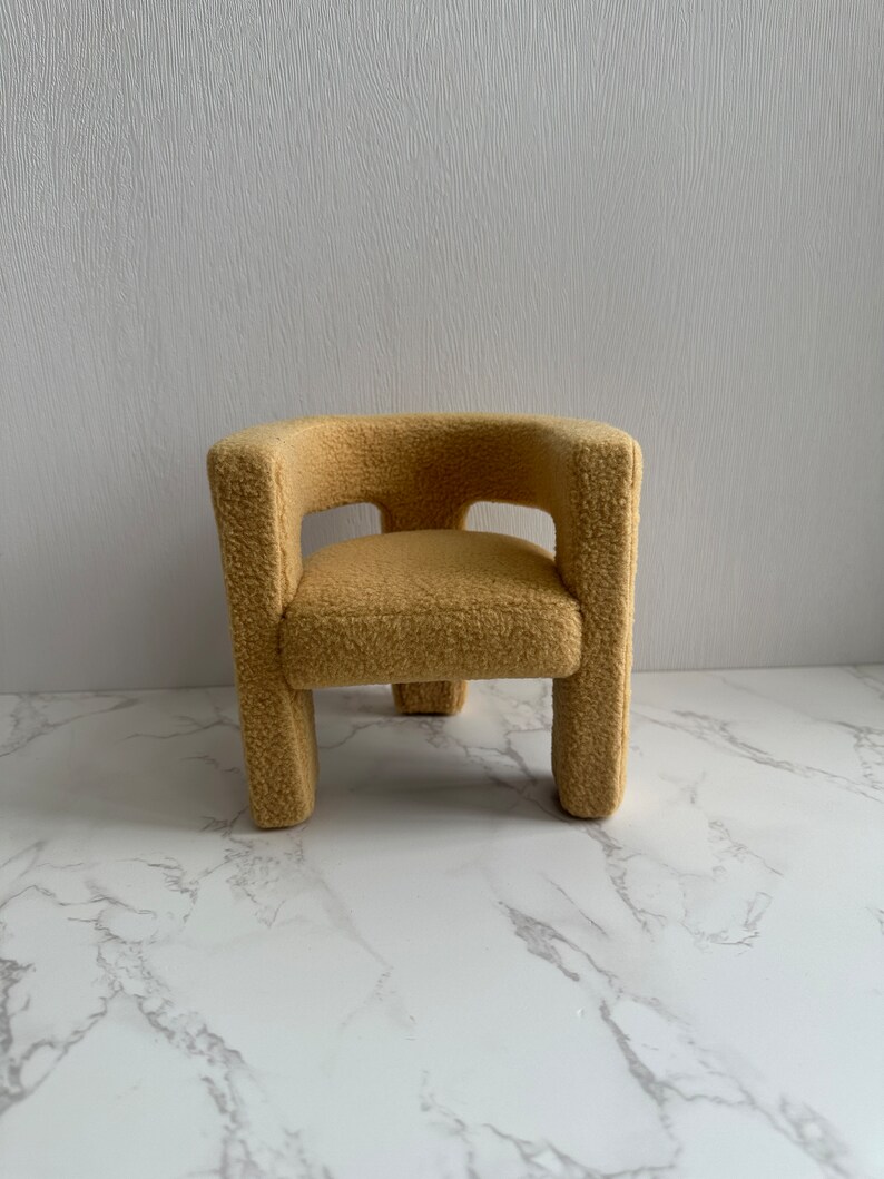 May include: A small, plush armchair with a rounded back and a light brown color. The chair has a simple, modern design and is made of a soft, fuzzy material.