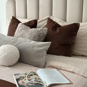 May include: A beige scalloped headboard behind a bed with striped beige bedding, two dark brown square pillows, a textured gray rectangular pillow, a small white round textured pillow, and an open book with a string of white beads resting on top.