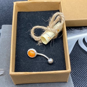 Könnte beinhalten: Eine kleine braune Pappbox mit schwarzem Innenraum. In der Box befindet sich ein silberner Bauchnabelpiercing mit einem orangefarbenen Stein. Die Box ist mit Schnur gebunden und ein kleines, zusammengerolltes Papier befindet sich im Inneren.