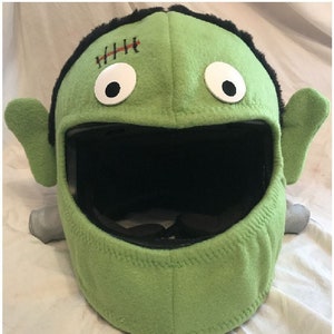 May include: Green fleece motorcycle helmet cover with a Frankenstein face, including stitched scar, googly eyes, and black mouth.