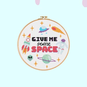 Puede incluir: Un aro de bordado blanco con un diseño de punto de cruz colorido que presenta un cohete, un OVNI, un cometa, un astronauta y el texto "GIVE ME some SPACE".