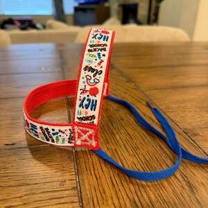 May include: A red and white ribbon with the text "Back to School" and other school-themed graphics. The ribbon is attached to a blue cord, likely for a backpack or other school item. The ribbon is on a wooden surface.