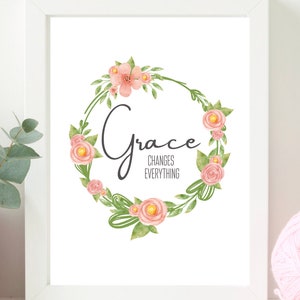 May include: A white framed print featuring a floral wreath design. The wreath is composed of pink flowers and green leaves. The word "Grace" is written in black cursive script within the wreath. The text "Changes Everything" is written in black below the word "Grace".