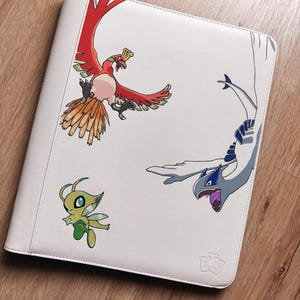 Puede incluir: Una carpeta blanca con un gráfico impreso de tres criaturas: una figura de ave roja y naranja, una figura de dragón azul claro y blanca, y una pequeña figura de hada verde. La carpeta tiene un acabado liso y un pequeño logotipo en relieve.