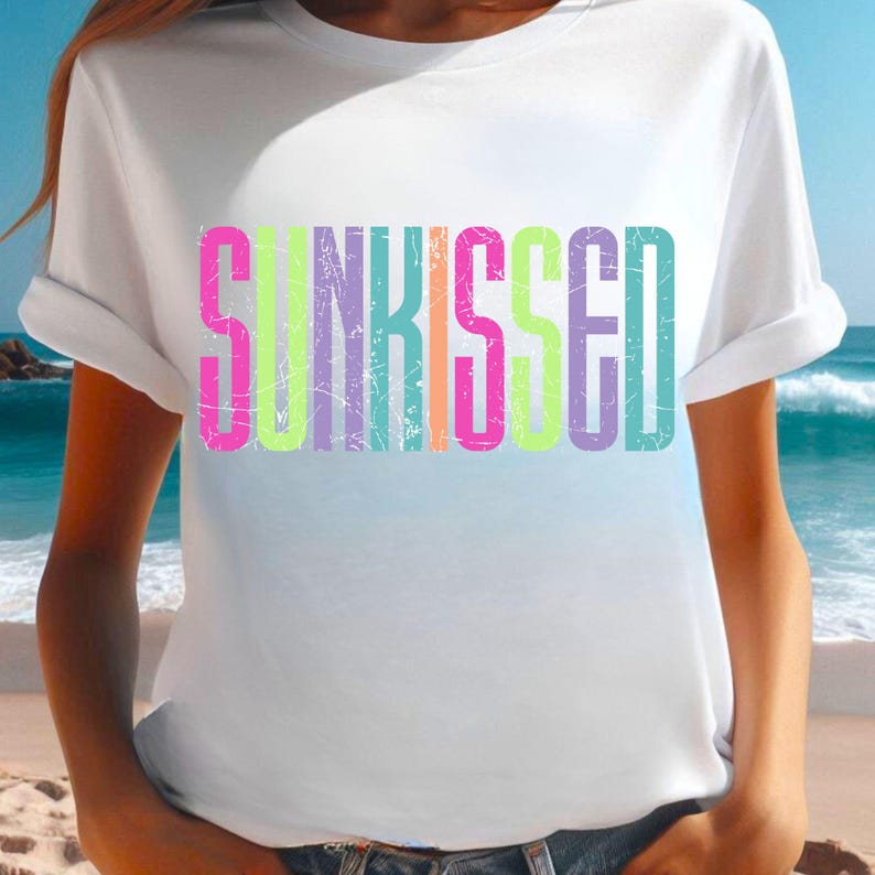 May include: White t-shirt with the word "SUNKISSED" in colorful, distressed lettering. The text features a gradient of pink, yellow, green, and blue hues. The shirt has rolled-up sleeves and is set against a beach background.