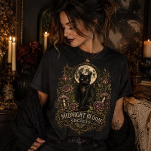 Pode incluir: Uma t-shirt de manga curta cinzento-carv&atilde;o com uma impress&atilde;o gr&aacute;fica de um gato preto sentado perante uma lua cheia, rodeado por rosas e cogumelos. O texto "MIDNIGHT BLOOM SOCIETY" est&aacute; impresso numa fonte dourada decorativa.