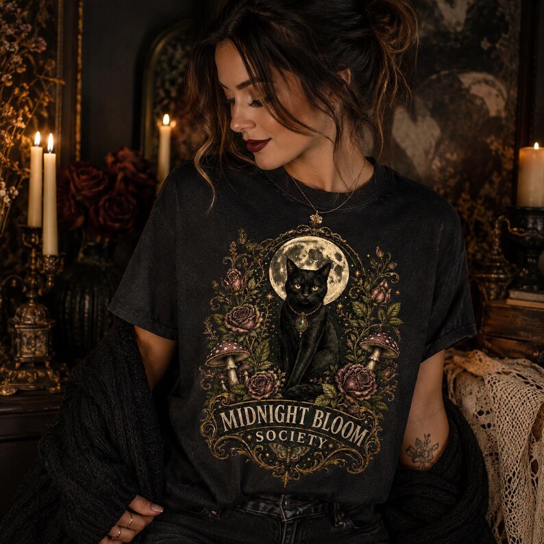 Pode incluir: Uma t-shirt de manga curta cinzento-carv&atilde;o com uma impress&atilde;o gr&aacute;fica de um gato preto sentado perante uma lua cheia, rodeado por rosas e cogumelos. O texto "MIDNIGHT BLOOM SOCIETY" est&aacute; impresso numa fonte dourada decorativa.