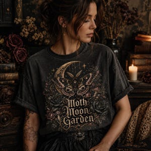 Pode incluir: Uma t-shirt cinza-carv&atilde;o com uma estampa gr&aacute;fica de uma mariposa, uma lua crescente, rosas e cogumelos. O texto "Moth Moon Garden" est&aacute; impresso em uma fonte decorativa abaixo da mariposa. A camisa tem gola redonda e mangas curtas.
