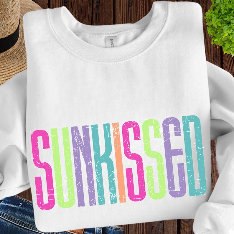 May include: White sweatshirt with the word "SUNKISSED" in large, distressed, multi-colored letters across the front. The letters are pink, green, purple, orange, and blue. The sweatshirt has a crew neck.