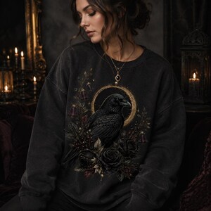 Woman wearing an oversized charcoal sweatshirt featuring the raven, gold eclipse and dark botanical graphic.