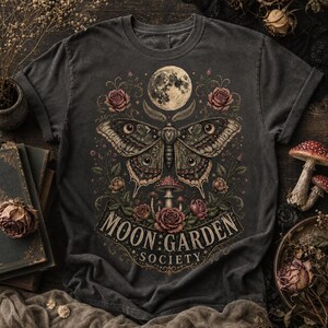 May include: A charcoal gray t-shirt featuring a graphic print of a large moth centered below a full moon. The design includes surrounding roses, small mushrooms, and a banner at the bottom with the text MOON GARDEN SOCIETY in a serif font.