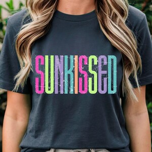 May include: Dark gray t-shirt with the word "SUNKISSED" in large, colorful, distressed lettering. The text features a gradient of pink, yellow, green, blue, and purple. The shirt is a casual style.