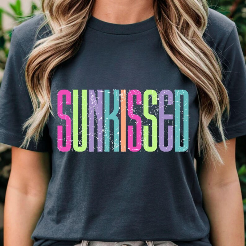 May include: Dark gray t-shirt with the word "SUNKISSED" in large, colorful, distressed lettering. The text features a gradient of pink, yellow, green, blue, and purple. The shirt is a casual style.