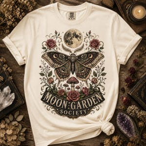 May include: A cream-colored short-sleeve t-shirt featuring a central graphic of a large moth, a full moon, roses, and mushrooms. Below the illustration, the text reads MOON GARDEN SOCIETY in a decorative black banner. The shirt has a crew neckline.