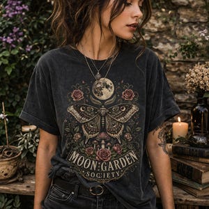 May include: A charcoal gray short-sleeve t-shirt featuring a graphic print of a large moth, a full moon, roses, and mushrooms. The text "MOON GARDEN SOCIETY" is printed in a decorative font at the bottom of the design. The shirt has a crew neckline.