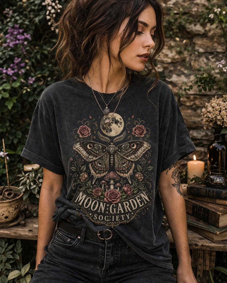 May include: A charcoal gray short-sleeve t-shirt featuring a graphic print of a large moth, a full moon, roses, and mushrooms. The text "MOON GARDEN SOCIETY" is printed in a decorative font at the bottom of the design. The shirt has a crew neckline.
