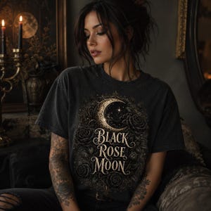 Pode incluir: Uma t-shirt cinzenta-carv&atilde;o de manga curta com uma impress&atilde;o gr&aacute;fica de uma lua crescente rodeada por rosas e folhagem detalhadas. O texto "Black Rose Moon" est&aacute; impresso numa fonte serifada decorativa no centro do design.
