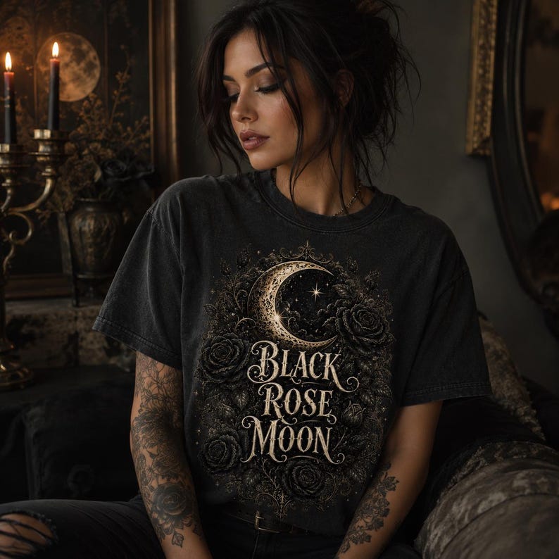 Pode incluir: Uma t-shirt cinzenta-carv&atilde;o de manga curta com uma impress&atilde;o gr&aacute;fica de uma lua crescente rodeada por rosas e folhagem detalhadas. O texto "Black Rose Moon" est&aacute; impresso numa fonte serifada decorativa no centro do design.