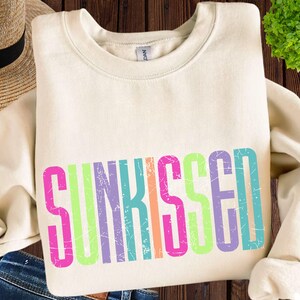 May include: Cream-colored sweatshirt with the word "SUNKISSED" in a colorful, distressed font. The text features pink, green, orange, purple, and blue letters. The sweatshirt is folded on a wooden surface.